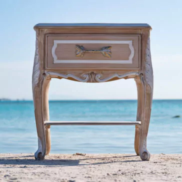 Ornate Italian Bedside Drawers An exquisite vintage wooden nightstand with elegantly carved legs and a drawer front, adorned with a brass handle in a leafy design, stands majestically on a sandy beach against the backdrop of clear blue waters and the distant horizon. This beautiful piece perfectly captures the charm of our Ornate Italian Bedside Drawers.