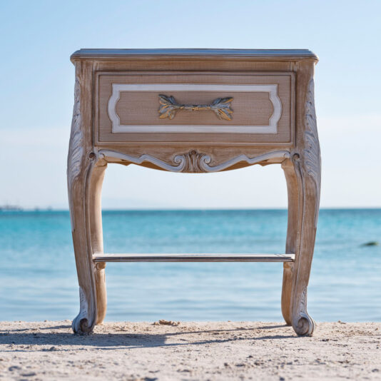 An exquisite vintage wooden nightstand with elegantly carved legs and a drawer front, adorned with a brass handle in a leafy design, stands majestically on a sandy beach against the backdrop of clear blue waters and the distant horizon. This beautiful piece perfectly captures the charm of our Ornate Italian Bedside Drawers.