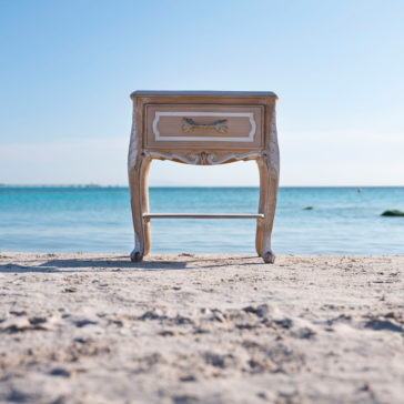 Ornate Italian Bedside Drawers An Ornate Italian Bedside Drawers nightstand stands on a sandy beach with a calm blue sea in the background. The sky is clear, and the horizon is visible, creating a serene and surreal scene.