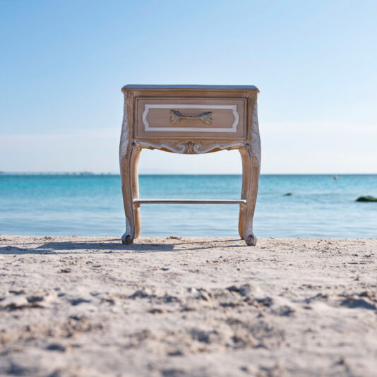 An Ornate Italian Bedside Drawers nightstand stands on a sandy beach with a calm blue sea in the background. The sky is clear, and the horizon is visible, creating a serene and surreal scene.