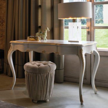 Ornate Italian Desk With Drawers An Ornate Italian Desk With Drawers, featuring elegant curved legs and a polished surface, stands by a window with flowing curtains. A cream-colored, tufted pouf is placed beneath the desk. A modern table lamp and decorative items, including a small green horse figurine, sit atop the desk.