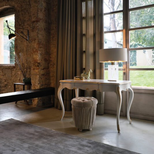 A stylish interior features the Ornate Italian Desk With Drawers, adorned with a lamp and a small decorative item, positioned in front of a large window. A plush round stool is placed underneath the desk. On the left side of the room, there is a brick wall decorated with a sconce and accompanied by a wooden bench.