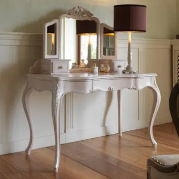 Ornate Italian Dressing Table The Ornate Italian Dressing Table, with its curved legs and intricate carvings, is painted in a light pastel color. This elegant piece features a tri-fold mirror, two small drawers on each side, and a central drawer. Topped with a lamp featuring a dark shade and various perfumes arranged on its surface, it epitomizes vintage elegance.