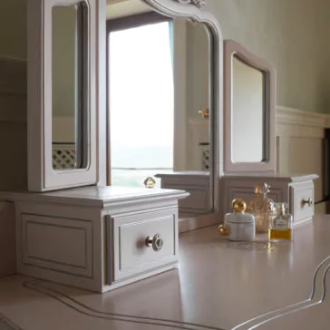 Ornate Italian Dressing Table A close-up shot of the Ornate Italian Dressing Table showcases its large central mirror flanked by two side mirrors. The table, with a light beige finish and silver accents, features small drawers. Decorative bottles and a small dish are arranged on the surface, and a window is visible in the background.