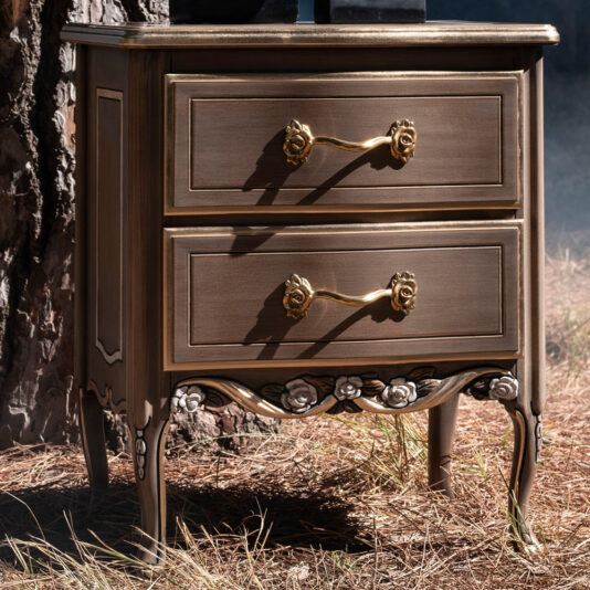 Ornate Louis Style Bedside Cabinet An elegant and compact Ornate Louis Style Bedside Cabinet, featuring two drawers adorned with golden handles. The piece is beautifully designed with intricate floral carvings at the bottom and is showcased outdoors on dry grass beside a tree trunk.