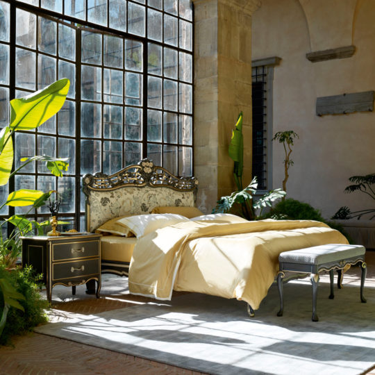 A luxurious bedroom showcases the Ornate Upholstered Italian Bed adorned with light beige bedding and complemented by a matching vintage nightstand. The room is illuminated by natural light streaming through large, industrial-style windows, while lush green plants bring a touch of nature into the space.