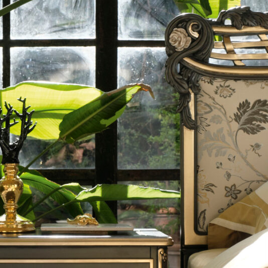 An Ornate Upholstered Italian Bed featuring a patterned headboard is positioned beside a window. Large green leaves of indoor plants are visible through the glass. On the nightstand beside the bed are a unique black tree-shaped sculpture and a golden lamp.