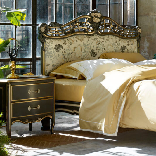 A luxurious bedroom features the Ornate Upholstered Italian Bed, characterized by its intricate floral-patterned headboard and elegant gold trim. The bed is dressed in beige and white bedding. Beside it, there's a matching nightstand adorned with a golden candle holder and decorative items. Large windows bring in natural light.