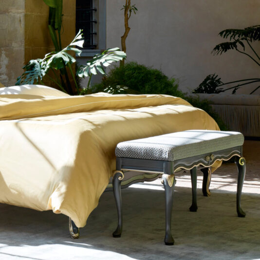 A sunny bedroom featuring the Ornate Upholstered Italian Bed adorned with light yellow bedding and a grey bench at its foot. The room is accentuated by large green plants, a window with light streaming in, and walls in neutral tones, creating a serene and inviting atmosphere.