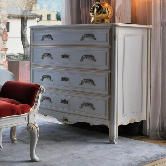 A vintage white dresser with ornate silver handles stands on a light rug, set against a backdrop of semi-transparent curtains. Nearby is the Tall Louis Style Chest Of Drawers. A red cushioned bench with white wooden legs adds a pop of color. On top of the dresser is a shiny golden decorative object.