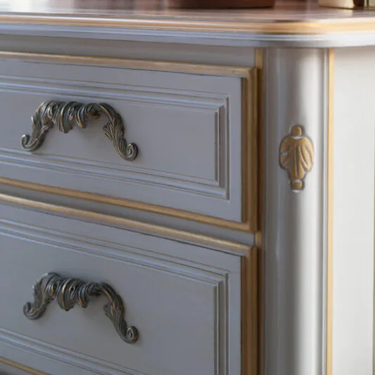 Tall Louis Style Chest Of Drawers Close-up of the Tall Louis Style Chest Of Drawers featuring ornate brass handles and decorative carvings. The dresser is painted in a light color with gold accents, which highlight the intricate details and craftsmanship.