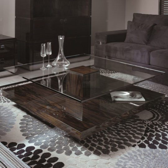 A modern living room featuring the Tiered Tempered Glass Top Coffee Table with a wooden base. On the table are a decanter, two champagne glasses, and a magazine. The room is furnished with a dark upholstered sofa and a patterned area rug, complemented by a dark wooden cabinet in the background.