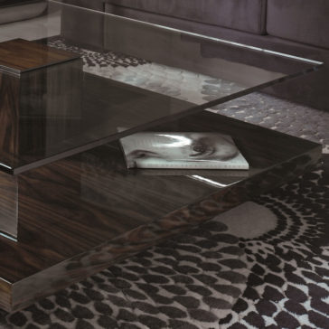 The Tiered Tempered Glass Top Coffee Table, featuring a wooden base and a modern design, is showcased with a magazine resting on its lower wooden shelf. The floor underneath boasts a patterned rug in shades of gray, while a portion of the sofa peeks from the background.
