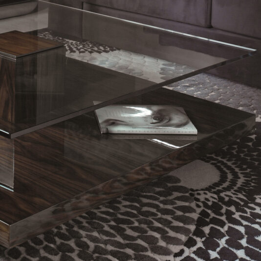Tiered Tempered Glass Top Coffee Table The Tiered Tempered Glass Top Coffee Table, featuring a wooden base and a modern design, is showcased with a magazine resting on its lower wooden shelf. The floor underneath boasts a patterned rug in shades of gray, while a portion of the sofa peeks from the background.