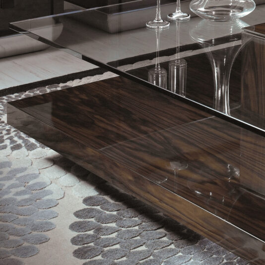 Tiered Tempered Glass Top Coffee Table The Tiered Tempered Glass Top Coffee Table with its sleek, dark wooden base stands on a plush, patterned rug. The surface reflects two elegant wine glasses and other minimally visible glassware, contributing to the chic and sophisticated ambiance of the room.