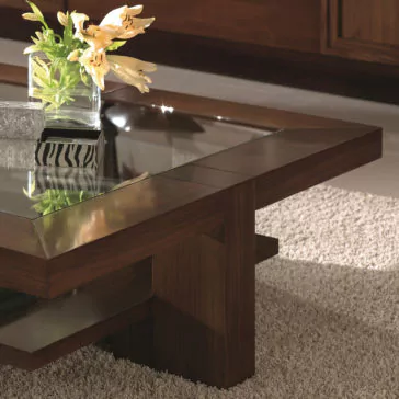 A Wooden Square Coffee Table With Glass Top sits on a light gray carpet. On the table, there's a clear glass vase containing yellow and white flowers.