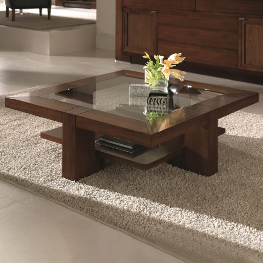 A Wooden Square Coffee Table With Glass Top and a lower shelf sits on a plush beige rug in a modern living room. The table holds a small decorative vase with yellow flowers and a striped rectangular object, possibly a small box or book. A wooden cabinet is in the background.