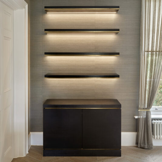 A minimalist interior showcases the Bespoke Cabinet And Floating Shelves — a dark wooden cabinet paired with four evenly spaced floating shelves above, all illuminated from underneath. The background wall features a subtle texture, and sheer curtains partly cover a window on the right side.
