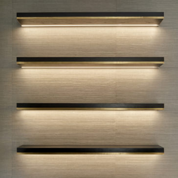 The image shows four horizontal, wall-mounted floating shelves from the Bespoke Cabinet And Floating Shelves collection, evenly spaced. These shelves feature a minimalistic design with a dark finish and built-in recessed lighting that beautifully illuminates the beige wall behind them, creating a modern and elegant appearance akin to custom cabinetry.
