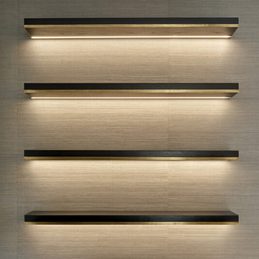 The image shows four horizontal, wall-mounted floating shelves from the Bespoke Cabinet And Floating Shelves collection, evenly spaced. These shelves feature a minimalistic design with a dark finish and built-in recessed lighting that beautifully illuminates the beige wall behind them, creating a modern and elegant appearance akin to custom cabinetry.