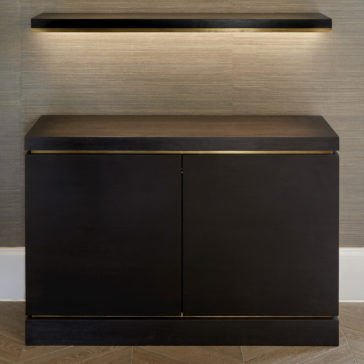 A Bespoke Cabinet And Floating Shelves in dark brown with gold trim stands against a textured beige wall. Above it, the matching floating shelves, complete with soft underlighting, add a modern touch. The floor showcases a light wood herringbone pattern.