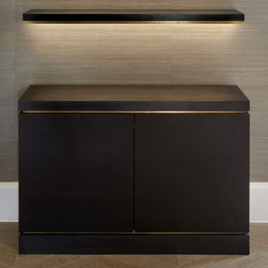 A Bespoke Cabinet And Floating Shelves in dark brown with gold trim stands against a textured beige wall. Above it, the matching floating shelves, complete with soft underlighting, add a modern touch. The floor showcases a light wood herringbone pattern.