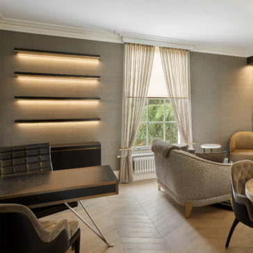 A stylish office space featuring a large wooden desk with a cushioned black chair, a beige-tiled floor, two cozy armchairs, a beige sofa, and a small round table. The Bespoke Cabinet And Floating Shelves add functionality to the room, while modern wall-mounted lights and natural light from elegant curtains provide ample illumination.