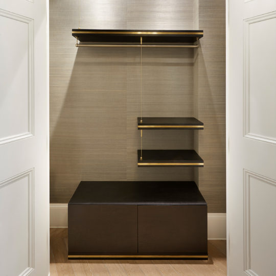 The Bespoke Hallway Storage system offers a spacious walk-in closet with an open door, showcasing a modern design. It features a dark wood lower cabinet accented with gold trim, a hanging rod, three floating shelves on the right, and a light gray textured wall background. Perfect for customized hallway storage solutions.