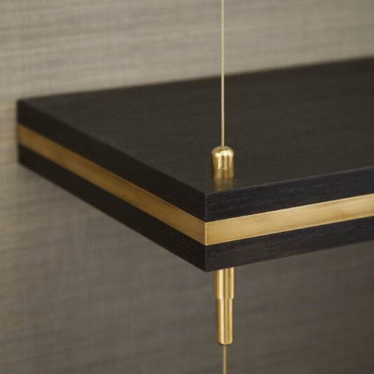 A close-up of Bespoke Hallway Storage, crafted from dark wood with a sleek brass accent. The shelf is elegantly suspended by a thin brass cable, showcasing its minimalist and modern design. In the background, a textured gray wall adds to the overall aesthetic.