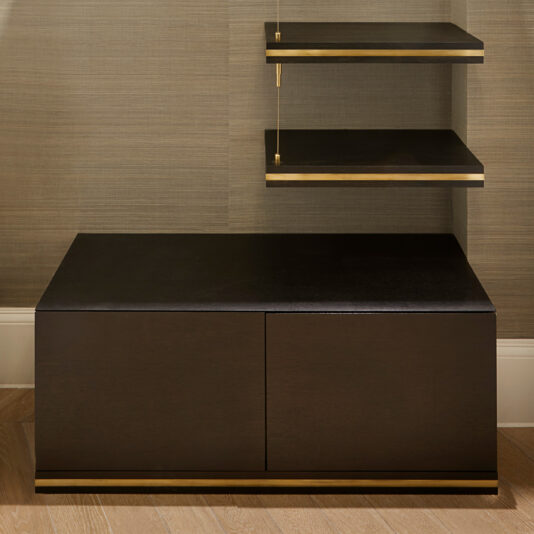 The Bespoke Hallway Storage unit boasts a dark finish with gold accents. It includes a minimalist rectangular base with closed compartments and two floating shelves above, both supported by gold brackets, all set against a neutral wall and hardwood floor.