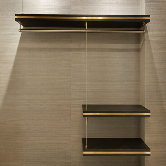 Introducing the Bespoke Hallway Storage: a modern closet with a minimalist design. It features two dark wooden shelves on the right side and a matching upper shelf with an attached hanging rod on the left, all supported by sleek metal brackets against a textured beige wall.