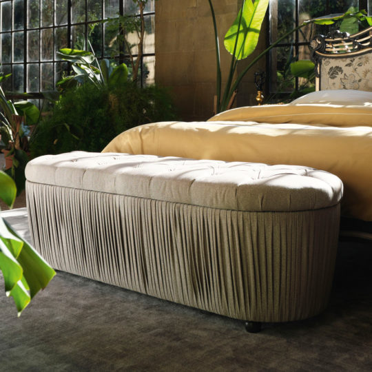 A luxurious bedroom features a Button Upholstered Ottoman Storage Bench at the foot of a bed with cream bedding. Large windows let in natural light, and potted plants add a green touch to the elegant space. The floor is covered with a dark area rug.