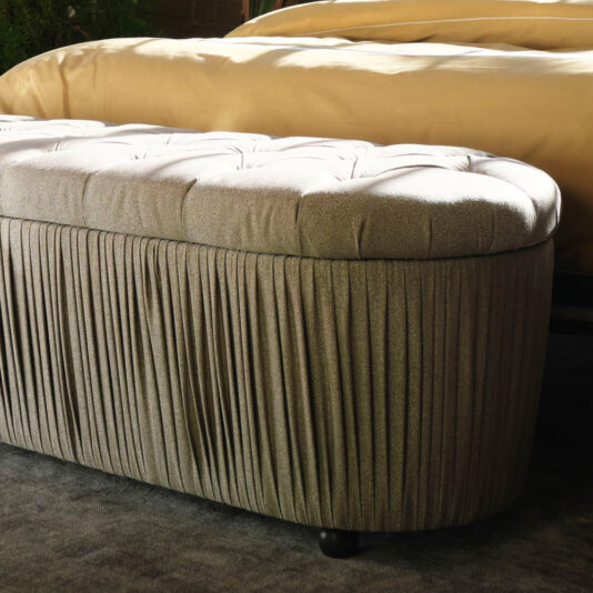 A Button Upholstered Ottoman Storage Bench, featuring an oval shape and a pleated fabric base, stands against the edge of a bed adorned with a beige blanket. The Italian designer piece boasts button upholstery that adds elegance to its plush, light-colored cushion, all nestled on top of a soft, dark carpet.
