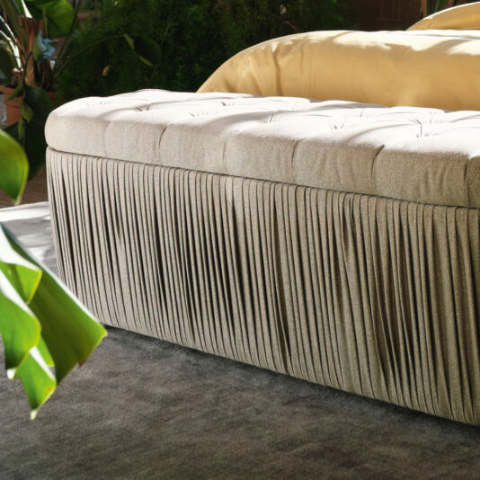 A plush, rectangular Button Upholstered Ottoman Storage Bench with a tufted, cushioned top and a pleated fabric base is placed on a soft, gray carpet. The button-upholstered ottoman is situated near a bed with golden bedding, and there are green plants in the background, adding a touch of natural decor.