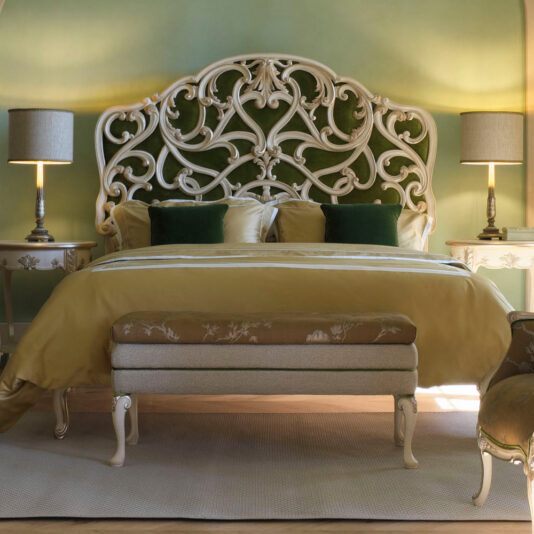 A luxurious bedroom featuring the Carved Rococo Style Bed with an ornate white wrought-iron tall headboard, adorned with green velvet accent pillows. Two matching bedside tables hold elegant lamps that cast soft, warm lighting. A neutral-toned bench sits gracefully at the foot of the bed.