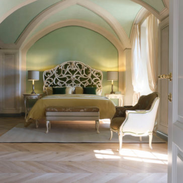 Carved Rococo Style Bed With Tall Headboard An elegant bedroom featuring a luxurious Carved Rococo Style Bed with a tall headboard, accompanied by two bedside tables with matching lamps. An upholstered armchair is placed by a sunlit window. The room is adorned in soft green and beige tones and boasts a curved, vaulted ceiling.