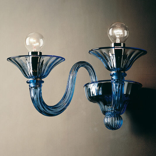The Classic Blue Glass Ribbed Detail Wall Light is a modern wall sconce that features classic blue glass with ribbed detailing. Each of its two arms gracefully curves outward from the base, supporting tulip-shaped holders for the light bulbs. The illuminated bulbs cast a warm glow, forming a symmetrical and elegant pattern on the surrounding wall.