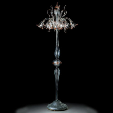 Classic Floral Detail Table Lamp The Classic Floral Detail Table Lamp boasts a tall and elegant design with a clear, ornate glass base. Its lampshade features intricate floral details that form flower shapes blooming outwards, adding a touch of timeless sophistication. The classic lamp is displayed against a dark background, accentuating its delicate craftsmanship.