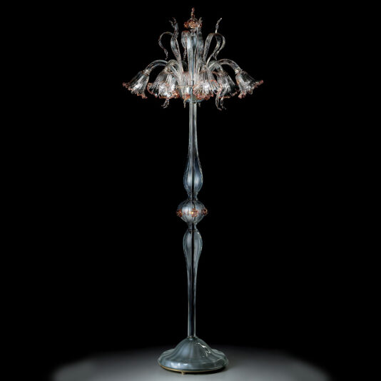 The Classic Floral Detail Table Lamp boasts a tall and elegant design with a clear, ornate glass base. Its lampshade features intricate floral details that form flower shapes blooming outwards, adding a touch of timeless sophistication. The classic lamp is displayed against a dark background, accentuating its delicate craftsmanship.