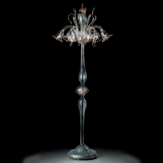 The Classic Floral Detail Table Lamp boasts a tall and elegant design with a clear, ornate glass base. Its lampshade features intricate floral details that form flower shapes blooming outwards, adding a touch of timeless sophistication. The classic lamp is displayed against a dark background, accentuating its delicate craftsmanship.