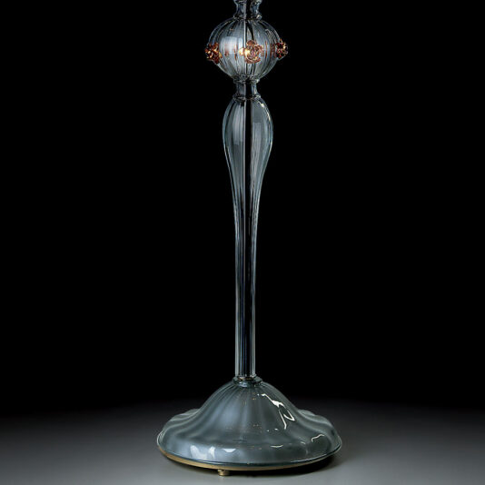 Classic Floral Detail Table Lamp The Classic Floral Detail Table Lamp is a tall, elegant glass fixture with a slender, elongated base that transitions into a spherical midsection adorned with classic floral details. This table lamp sits on a smooth, rounded base and is displayed against a dark background.