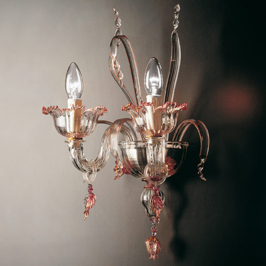 The Classic Floral Detail Wall Light is an ornate wall-mounted glass sconce with two candle-shaped bulbs that emit a warm glow. This elegant wall light features intricate glasswork with delicate, flowing lines and floral embellishments, adding a touch of sophistication to the décor. The background is a plain, dark wall.