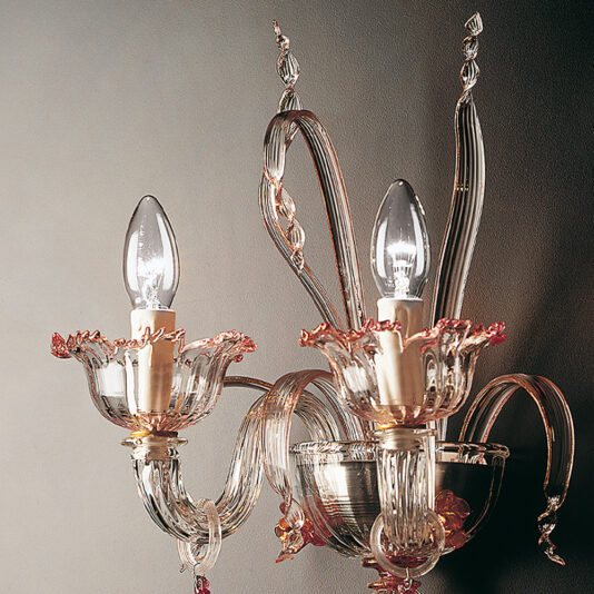 Classic Floral Detail Wall Light The Classic Floral Detail Wall Light is an ornate wall-mounted fixture adorned with two candle-shaped bulbs. It features intricate, curved designs crafted from clear glass and includes subtle pinkish accents on the flower-like holders for the bulbs, all set against a plain background.