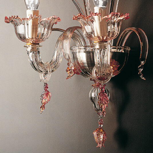 Classic Floral Detail Wall Light A close-up view of the Classic Floral Detail Wall Light reveals its intricately designed glass arms and classic floral motifs. The wall light features delicate pink and gold accents, two visible candle-shaped bulbs, and decorative elements hanging from its curved, transparent glass structure, reminiscent of a sophisticated chandelier.