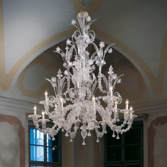 The Classic Glass Chandelier With Floral Detailing, showcasing an array of ornate arms, hangs from the ceiling of an elegantly decorated room with arched windows and intricate wall moldings. The chandelier is illuminated, casting a warm glow.