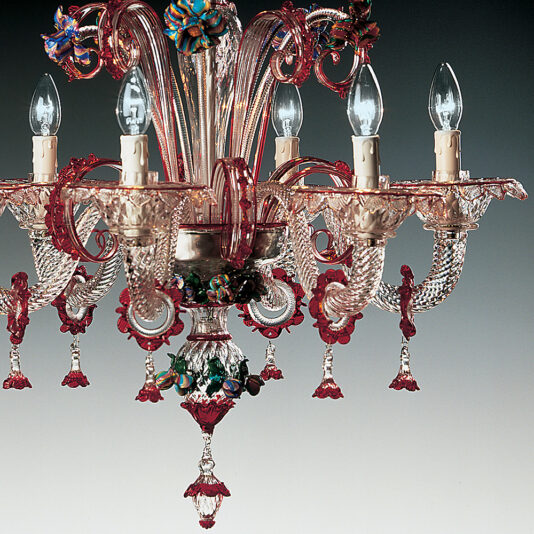 The Classic Glass Chandelier With Red Floral Detailing features intricate, swirling designs in shades of red and clear. This ornate chandelier has multiple arms, each holding a candle-shaped bulb. Decorative red floral elements and hanging glass drops enhance its elegance.