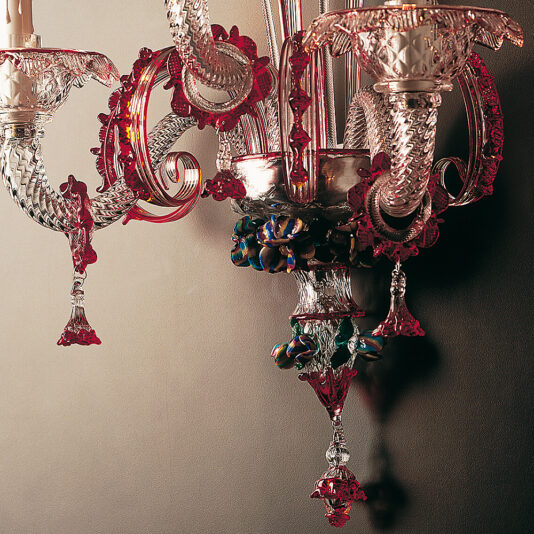 Classic Glass Wall Light With Red Floral Detailing A stunning Classic Glass Wall Light With Red Floral Detailing hangs against a wall. This ornate, vintage chandelier showcases intricate designs with clear and red floral accents, complete with candle-like extensions. The light is both elaborate and decorative, casting warm illumination and shadow patterns on the wall behind it.