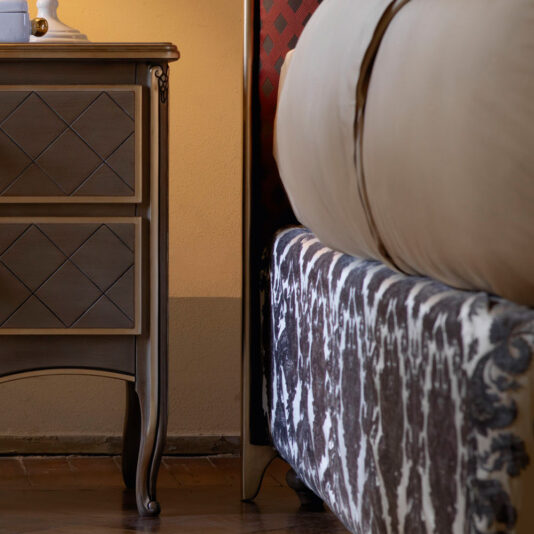 A close-up of a bedroom scene featuring the Classic Italian Bed with an upholstered frame and a wooden nightstand. The nightstand has a diamond pattern on its drawers and sits on wooden floors. The bedspread is light-colored, with a red accent wall behind.