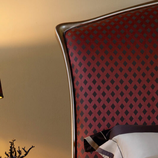 Close-up view of the Classic Italian Bed, featuring a padded headboard upholstered in a red and black diamond pattern fabric, accented with a bronze-colored frame. At the bottom of the image, a portion of a white pillow with dark edges can be seen. The warm beige wall behind enhances the classic appeal.
