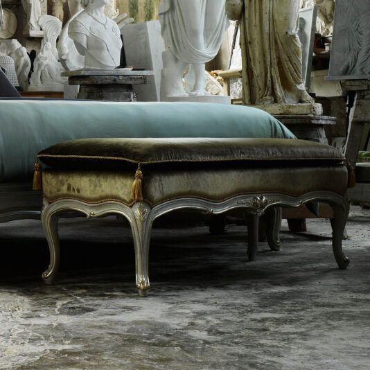 In an art studio filled with white classical statues and detailed sculptures, a vintage ottoman with dark upholstery and elegant wooden legs featuring gold tassels at the corners sits in front of a Classic Louis Style Bench upholstered in blue-green fabric. The floor is weathered.